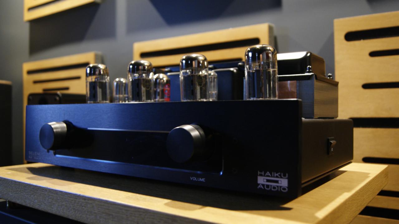 Czarny wzmacniacz lampowy Haiku Audio z widocznymi lampami i regulacją głośności, umieszczony na drewnianej półce na tle paneli akustycznych.