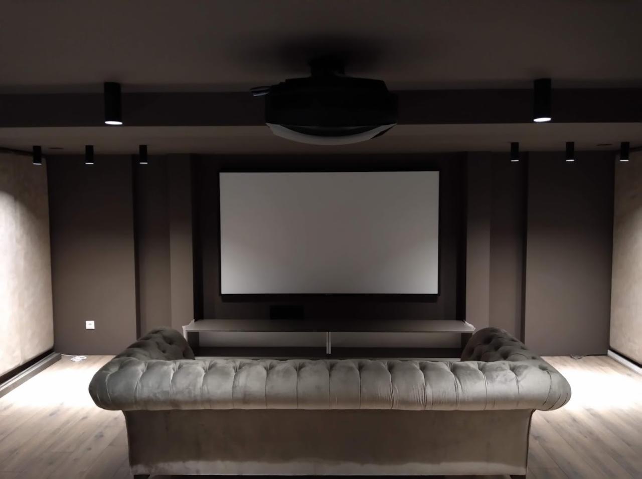 Domowe kino z projektorem sufitowym, ekranem projekcyjnym i szarą sofą w ciemnym pomieszczeniu z oświetleniem punktowym.