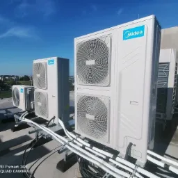 Instalacja kilku jednostek zewnętrznych klimatyzacji Midea na dachu budynku, widoczne białe rury instalacyjne i niebieskie logo producenta na tle błękitnego nieba.