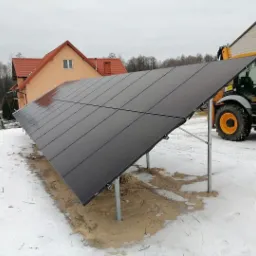 Rząd paneli słonecznych zamontowany na metalowych wspornikach na zasypanym śniegiem terenie, z domem jednorodzinnym i traktorem w tle.