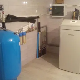 Instalacja kotłowni w Nowym Mieście nad Pilicą z niebieskim zbiornikiem ciśnieniowym, filtrami wody i kotłem grzewczym marki De Dietrich, widoczne rury i zawory.
