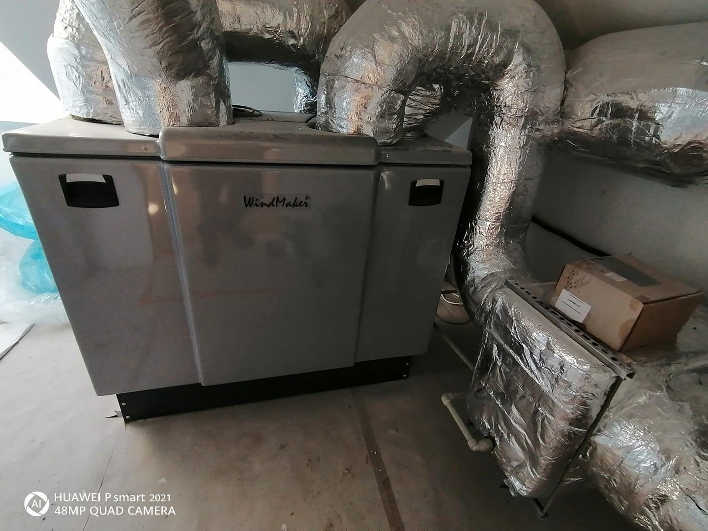 Szara centrala wentylacyjna WindMaker z podłączonymi, zaizolowanymi przewodami wentylacyjnymi w pomieszczeniu technicznym, widoczny karton na metalowej konstrukcji.