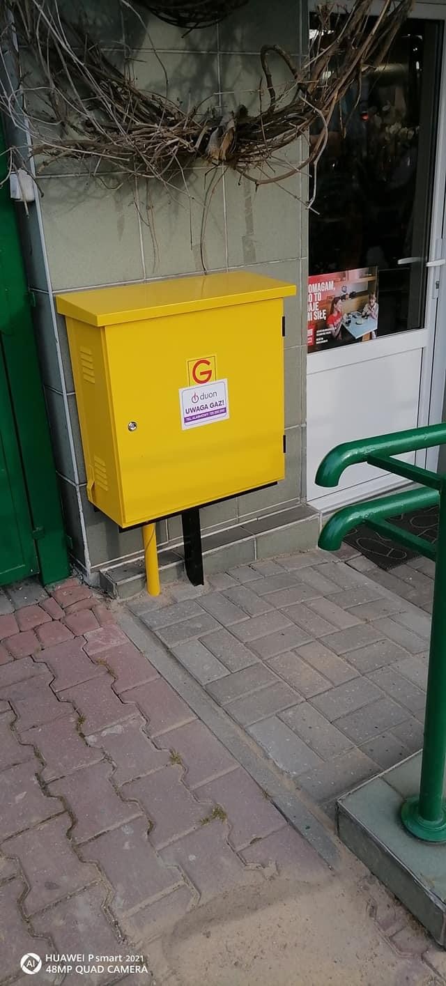 Żółta skrzynka gazowa z naklejką ostrzegawczą 'Uwaga Gaz!' zamontowana na ścianie budynku obok wejścia z zieloną barierką i dekoracją z gałęzi.