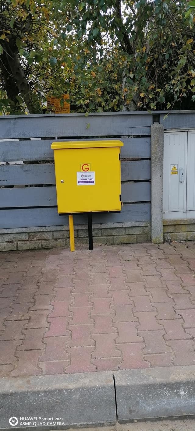 Żółta skrzynka gazowa zamontowana na zewnątrz budynku, z tabliczką ostrzegawczą 'Uwaga gaz!' i literą 'G', ustawiona na kostce brukowej, na tle szarego ogrodzenia i zieleni drzew.