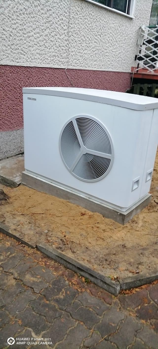 Biała pompa ciepła marki Stiebel Eltron zainstalowana na betonowej podstawie przed budynkiem z elewacją w kolorze kremowo-bordowym. Widoczna kostka brukowa i ziemia.