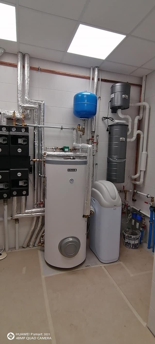 Biały bojler De Dietrich z niebieskim zbiornikiem wyrównawczym i szarą stacją uzdatniania wody, widoczne rury miedziane i izolowane, zainstalowane w pomieszczeniu z białymi płytkami ściennymi...