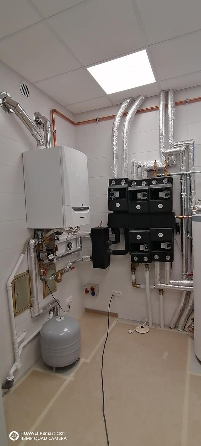 Nowoczesny, biały piec gazowy kondensacyjny zamontowany na ścianie z białymi płytkami, z widocznymi rurami miedzianymi i izolowanymi, srebrnymi rurami, oraz czarnymi modułami sterującymi...