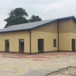 Parterowy budynek z beżową elewacją i ciemnymi oknami. Widoczne ułożone płyty chodnikowe i przygotowane materiały budowlane. Budynek o prostej architekturze z dwuspadowym dachem.