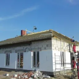 Budowa domu z białych bloczków: częściowo ocieplona ściana, robotnicy na drabinie montują elementy dachu. Widoczne rusztowania i gruz. Słoneczny dzień.