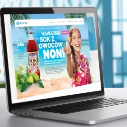 Laptop wyświetla stronę internetową z reklamą hawajskiego soku z owoców Noni, z uśmiechniętą kobietą w stroju hawajskim na tle plaży, butelką soku i owocami Noni, w tle biuro.