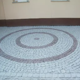 Ułożona kostka granitowa w kształcie spirali, z szarych i brązowych kamieni. Spirala tworzy wzór na podwórku przed budynkiem. Widoczne okna i rynna.