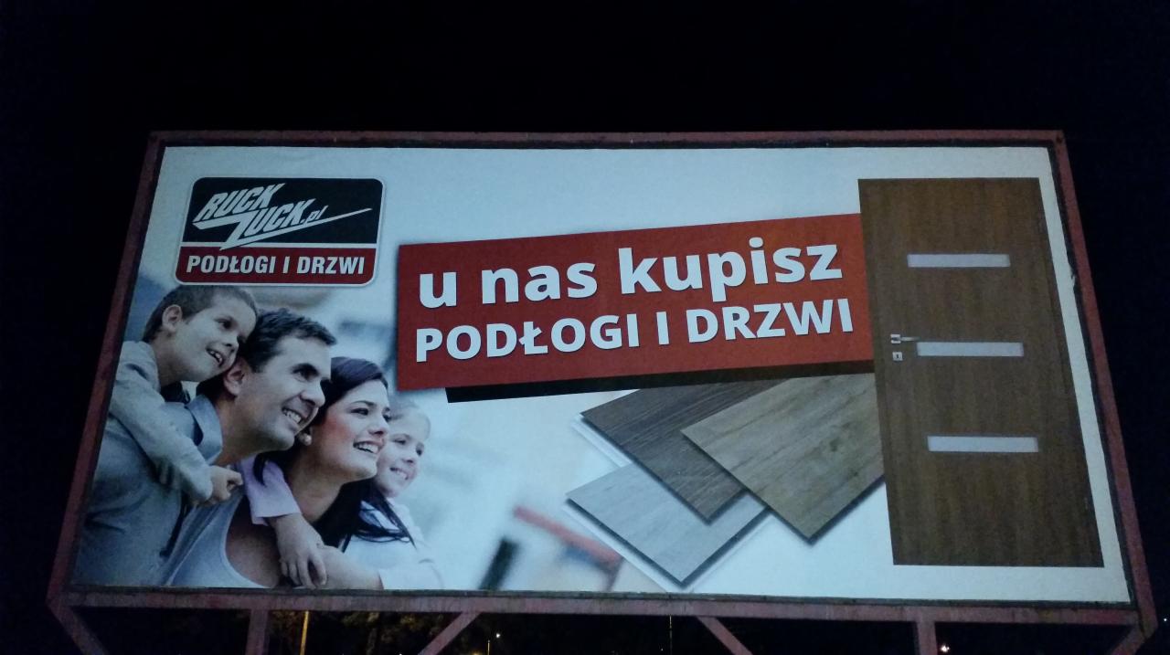 Billboard reklamowy firmy Ruck Zuck z hasłem 'U nas kupisz podłogi i drzwi', prezentujący uśmiechniętą rodzinę, próbki podłóg i brązowe drzwi.