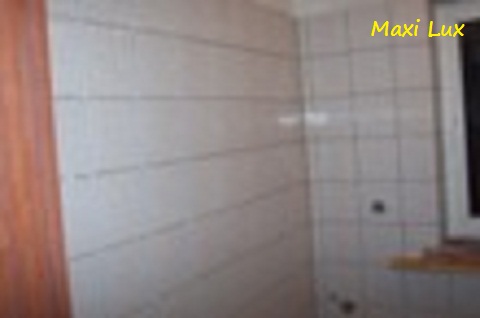 Fragment ściany łazienki z białymi, kwadratowymi płytkami. Widoczny fragment okna z parapetem i drzwi. Na górze żółty napis Maxi Lux.