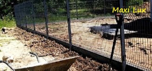 Montaż ogrodzenia panelowego z podmurówką betonową. Widoczna taczka i szpadel. Teren przygotowany pod instalację, z fragmentami ziemi. Na ogrodzeniu napis 'Maxi Lux'.