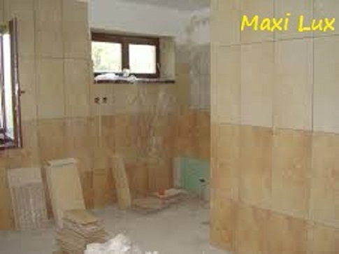 Wnętrze łazienki w trakcie remontu: częściowo wyłożone beżowe płytki na ścianach, okno, materiały budowlane na podłodze. Widoczny etap prac wykończeniowych.