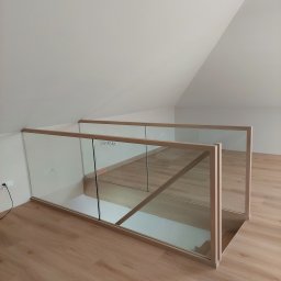 Schody Drewniane Bartosz Paczkowski - Szklana balustrada z drewnianą ramą zabezpieczająca schody, widok z góry na jasne drewniane panele podłogowe i białe ściany poddasza.