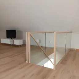 Nowoczesna balustrada szklana z drewnianymi słupkami na antresoli, widok na schody i salon z telewizorem.