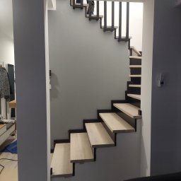 Schody Drewniane Bartosz Paczkowski - Schody drewniane z czarnymi wspornikami i metalowymi balustradami, widok z dołu, ściana w kolorze gołębim.