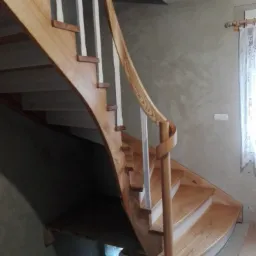 Zakręcone schody z jasnego drewna z białą balustradą, widok z dołu, naturalne oświetlenie, w tle ściana z oknem.