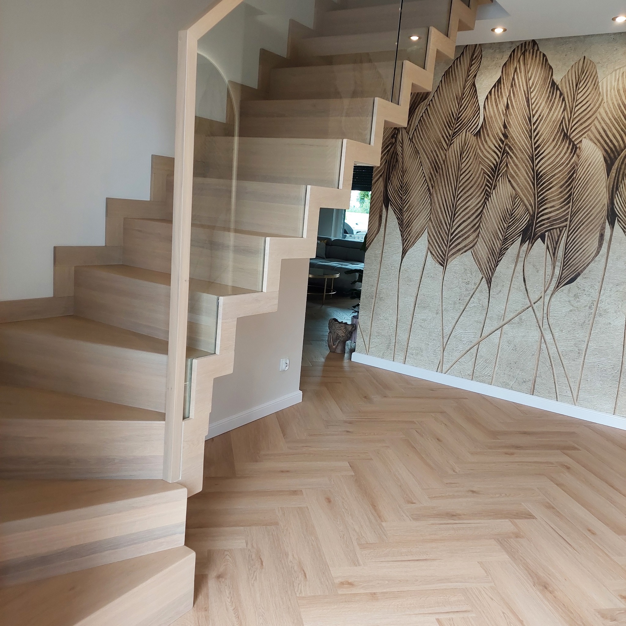 Nowoczesne, jasne schody drewniane z balustradą szklaną, parkiet w jodełkę na podłodze, ściana z dekoracyjną tapetą w liście.