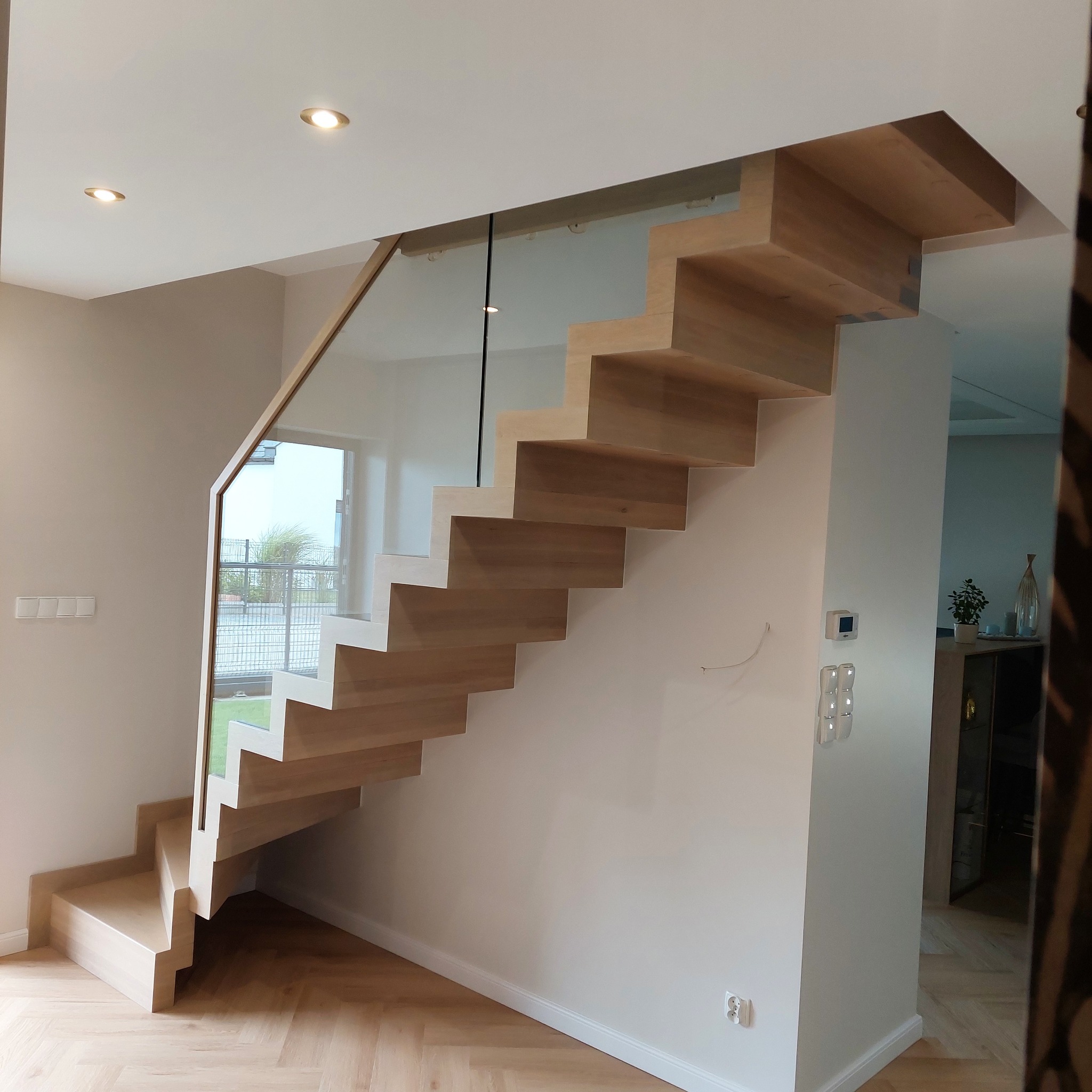 Nowoczesne, samonośne schody z jasnego drewna, z balustradą ze szkła hartowanego, widoczne z boku na tle jasnego wnętrza z parkietem w jodełkę.