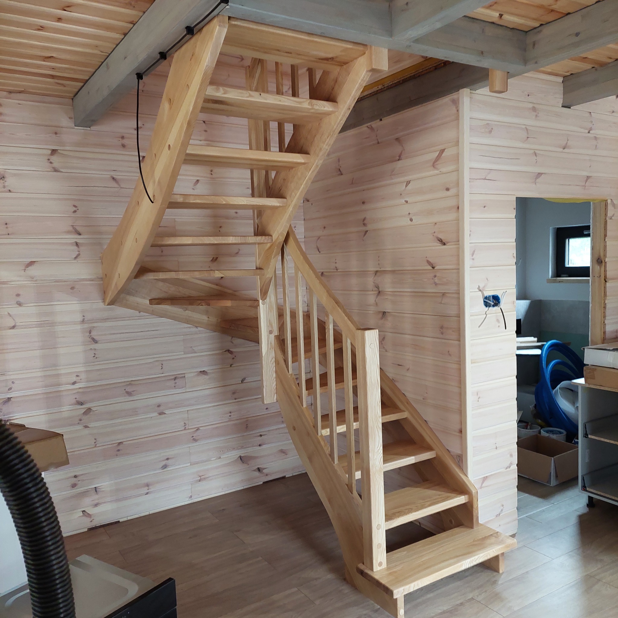 Jasne, drewniane schody z balustradą w surowym stanie, zamontowane w domu z boazerią, widoczny fragment podłogi i instalacja elektryczna w ścianie