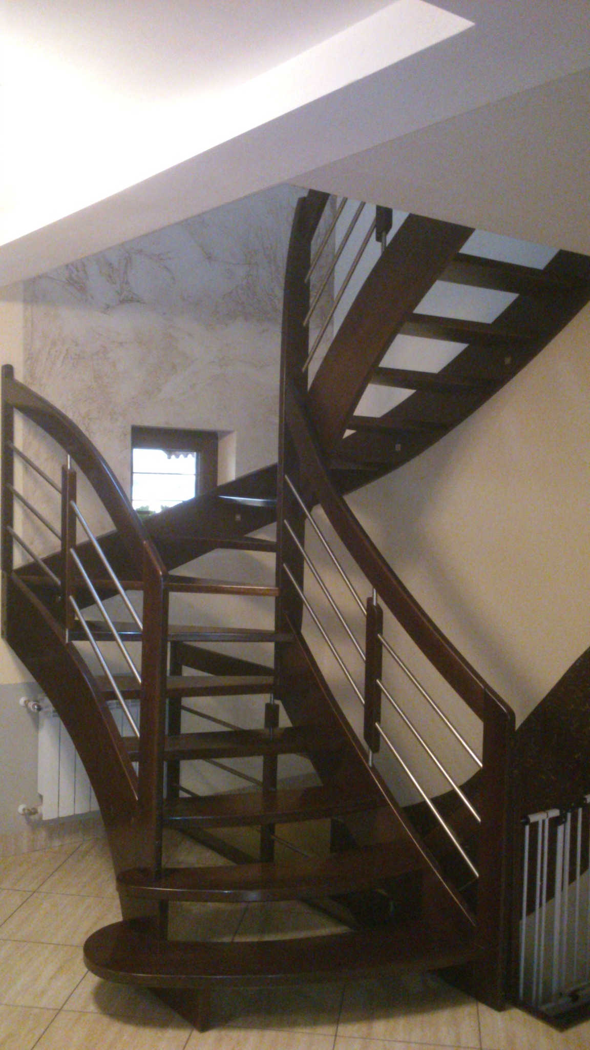 Spiralne schody drewniane z metalowymi poręczami wewnątrz pomieszczenia, widok z dołu, z oknem w tle.