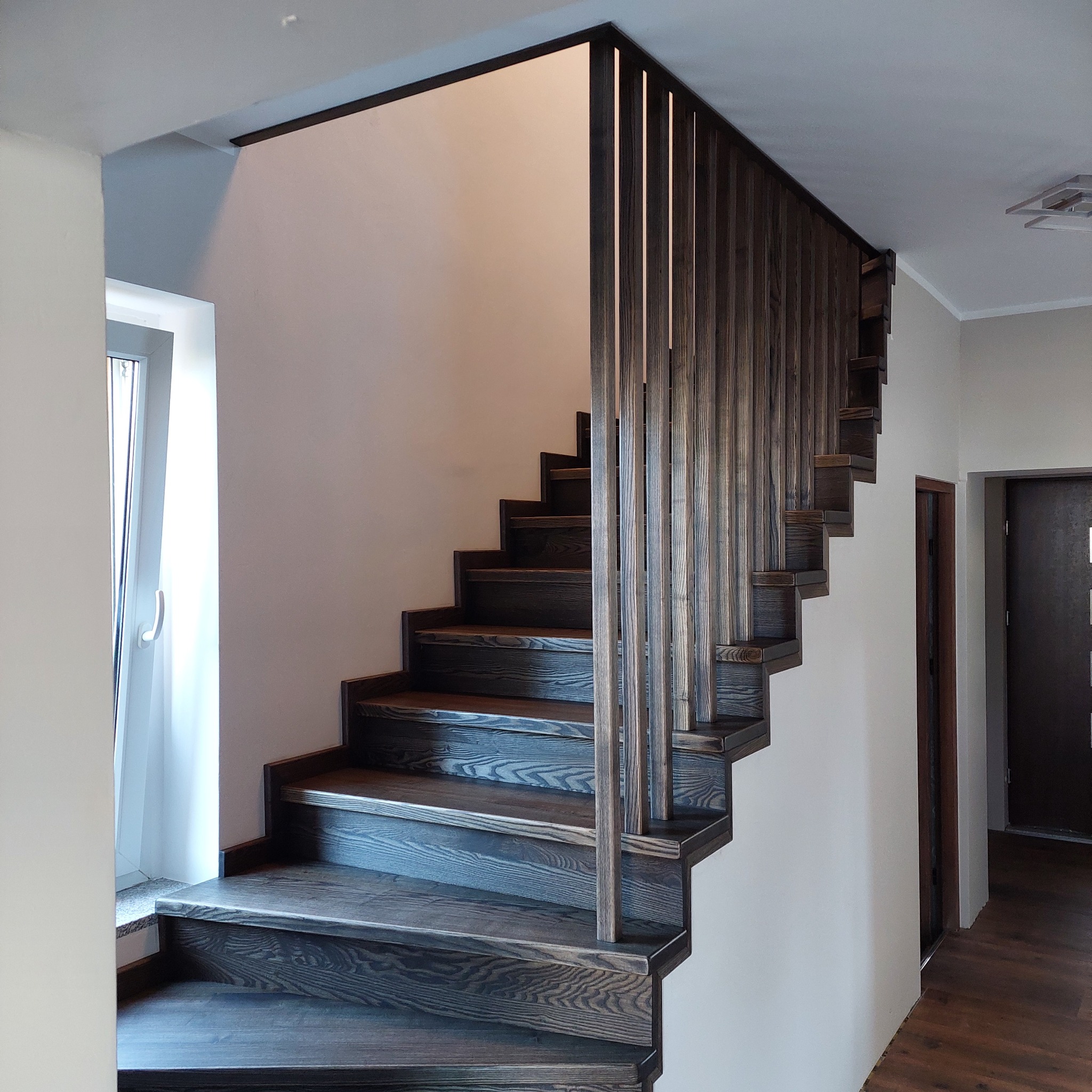 Ciemne, drewniane schody z pionową balustradą z desek, prowadzące do góry w jasnym wnętrzu.