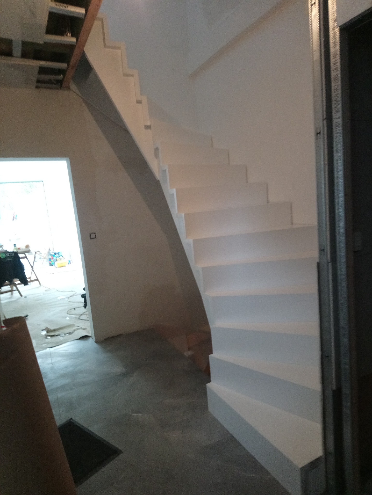 Białe, drewniane schody w trakcie montażu, widoczne elementy konstrukcyjne sufitu i ściany w stanie surowym, oraz narzędzia i materiały w tle.