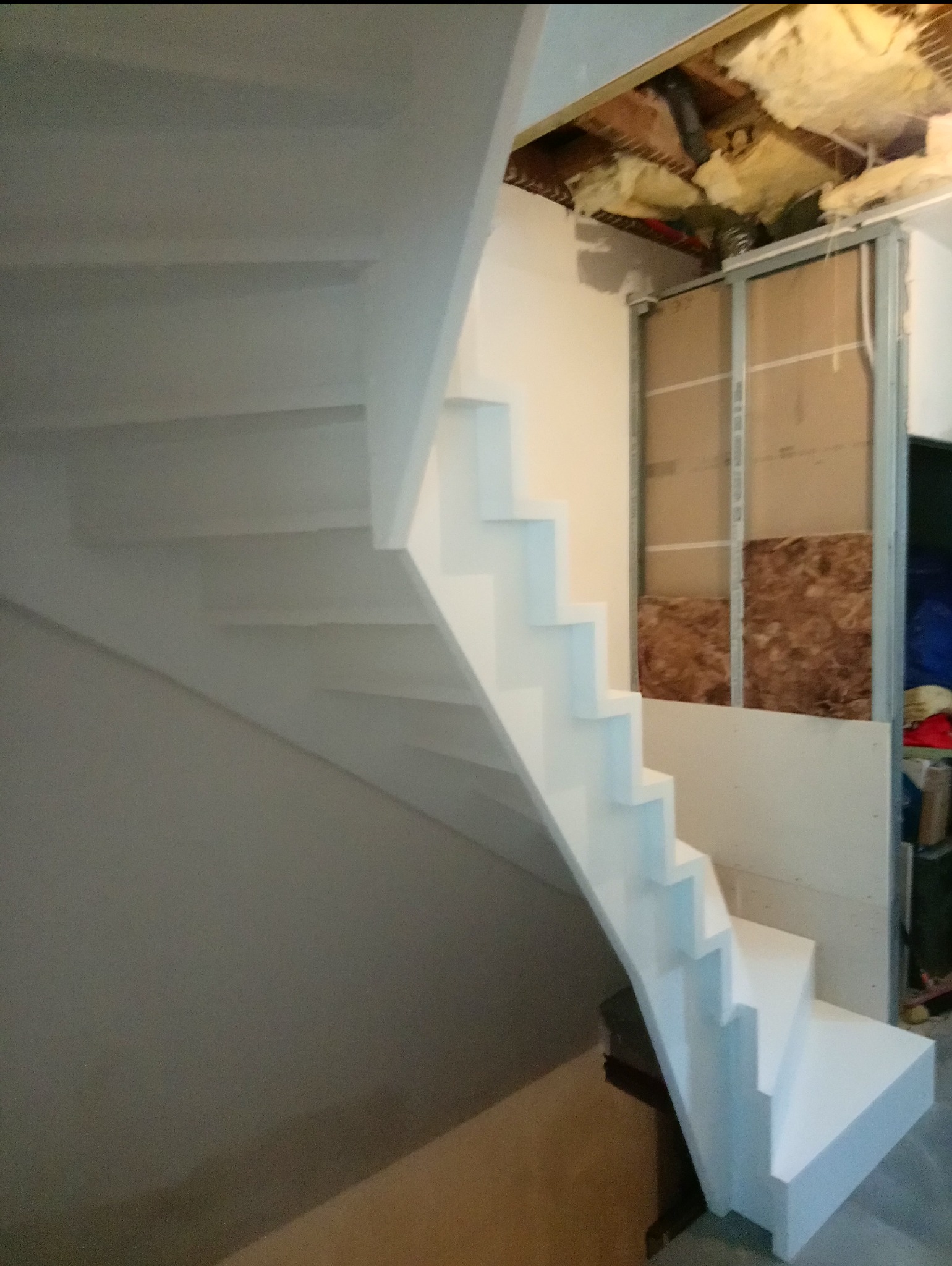 Białe schody w trakcie montażu, widoczne niedokończone wykończenia ścian i sufitu, izolacja termiczna w tle.