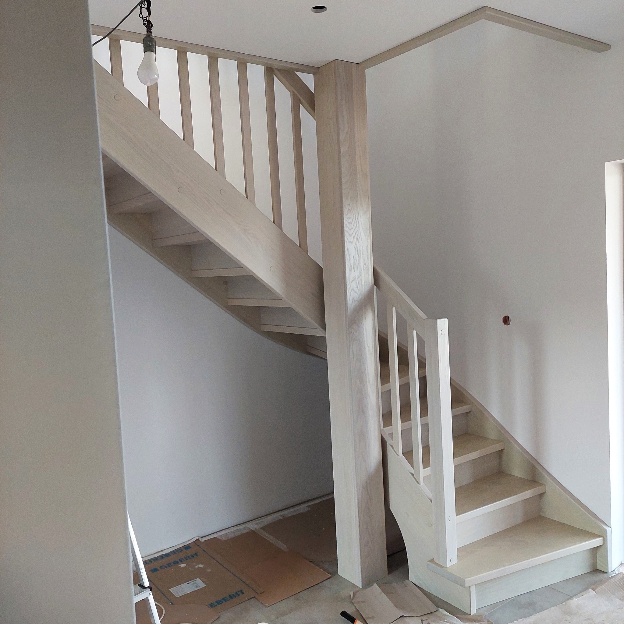 Jasne, drewniane schody z balustradą w trakcie montażu w nowym domu. Widoczne zabezpieczenia podłogi i surowa żarówka. Minimalistyczny design.