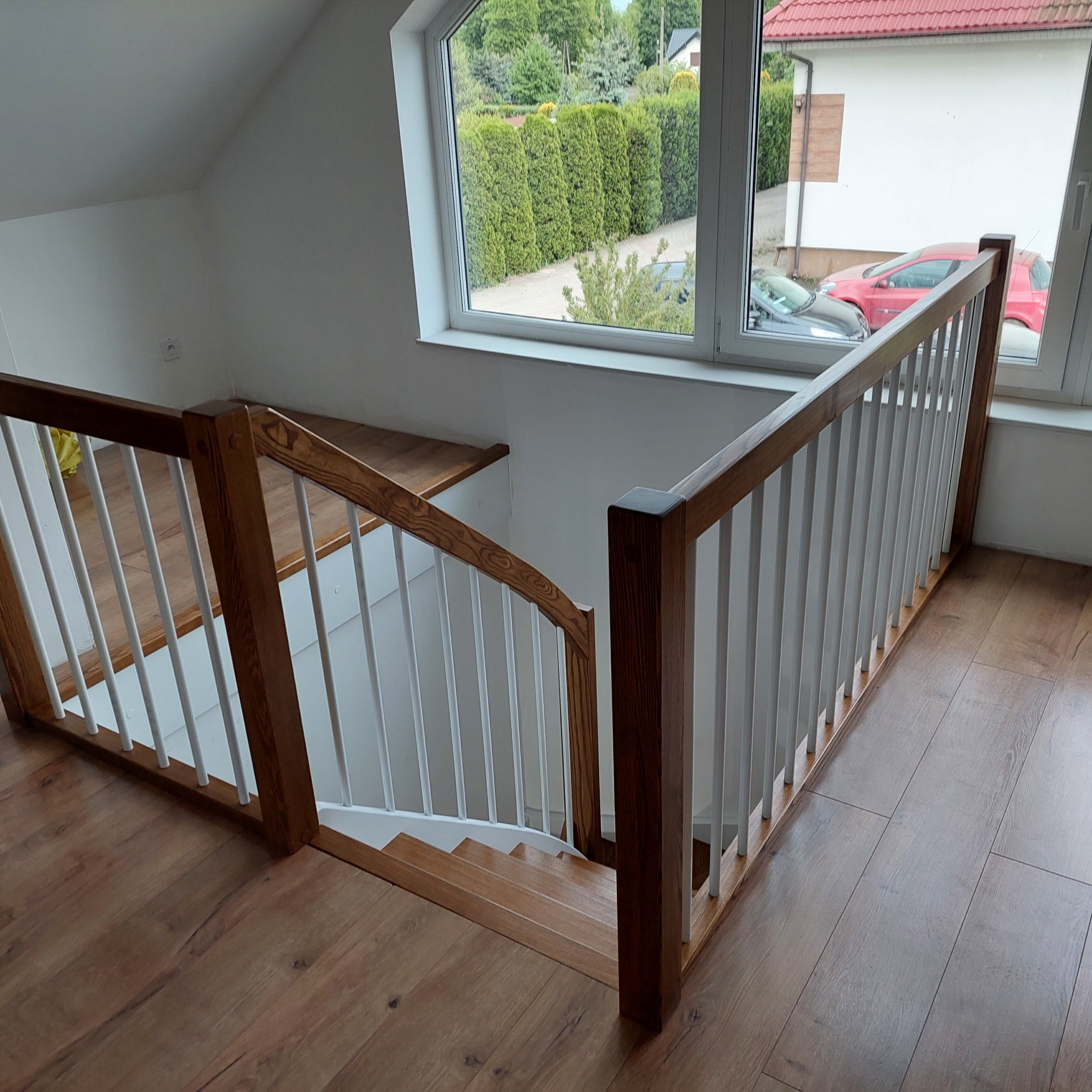 Kątowe ujęcie drewnianych schodów z białą balustradą w jasnym wnętrzu, widok z góry na dół, z oknem w tle i drewnianą podłogą.