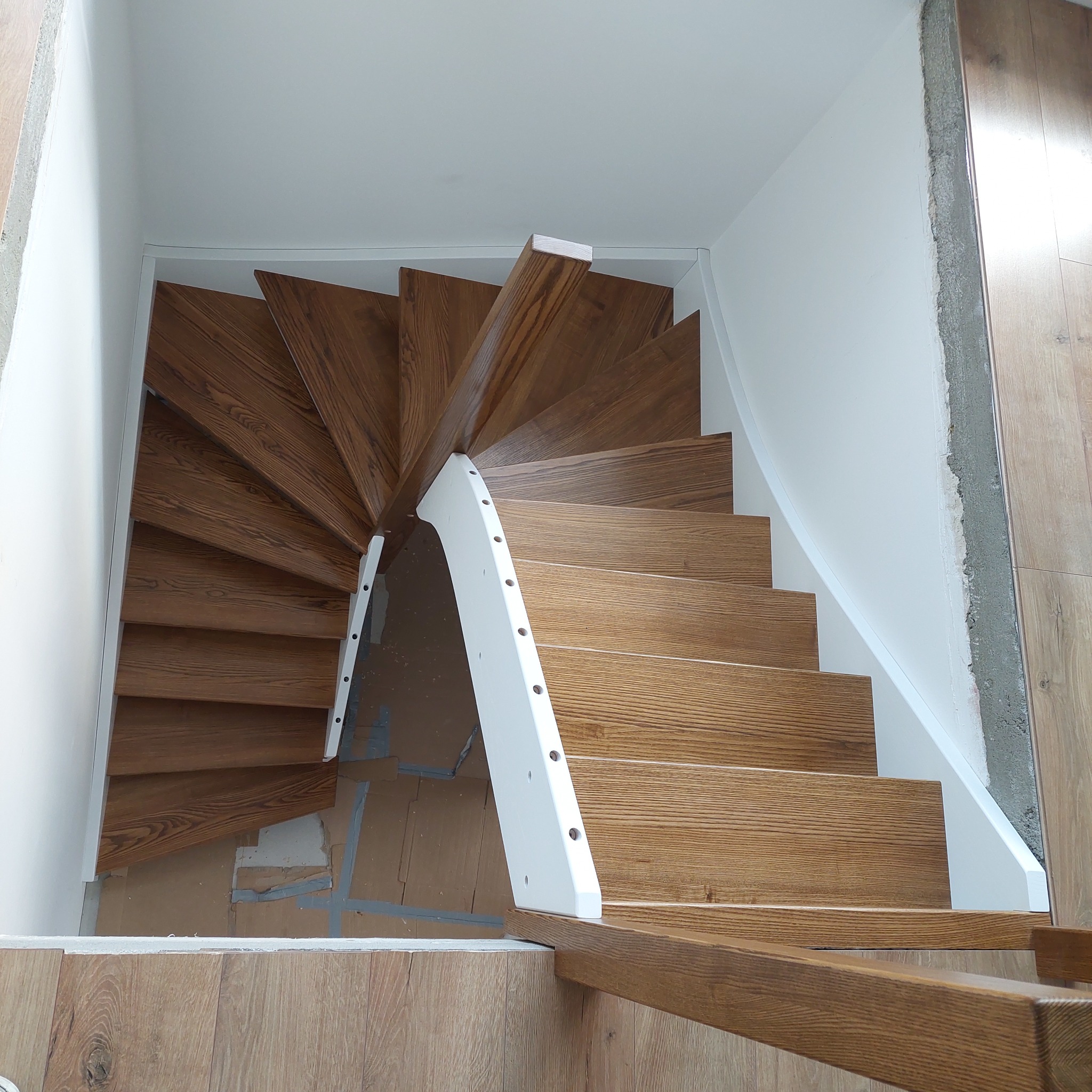 Spiralne schody drewniane z białymi balustradami w jasnym wnętrzu. Widok z góry ukazuje detale drewna i precyzję wykonania. Częściowo widoczne zabezpieczenia podłogi.