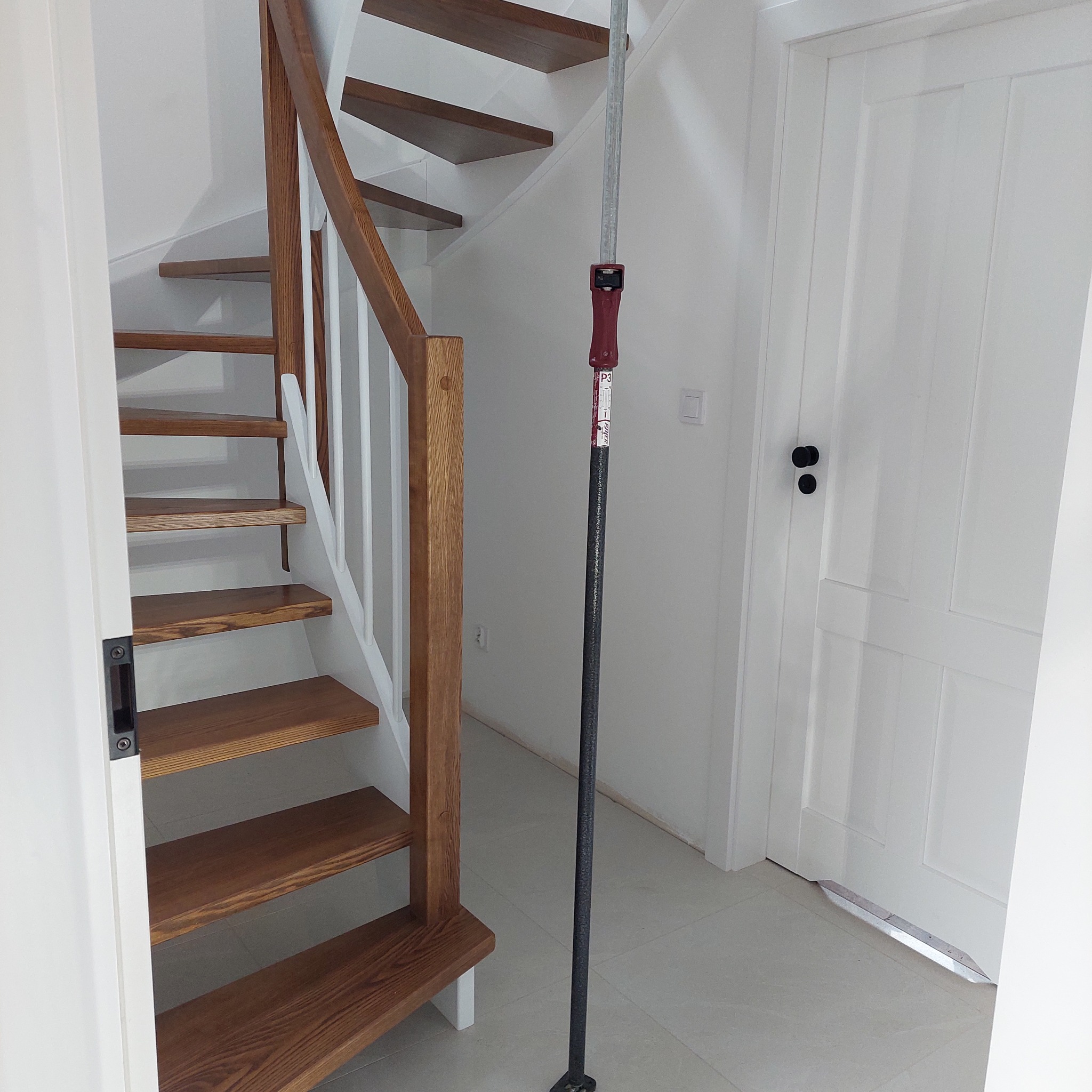 Jasne schody drewniane z białymi balustradami w nowoczesnym wnętrzu, widoczne narzędzie montażowe. Ujęcie z dołu, podkreślające konstrukcję.