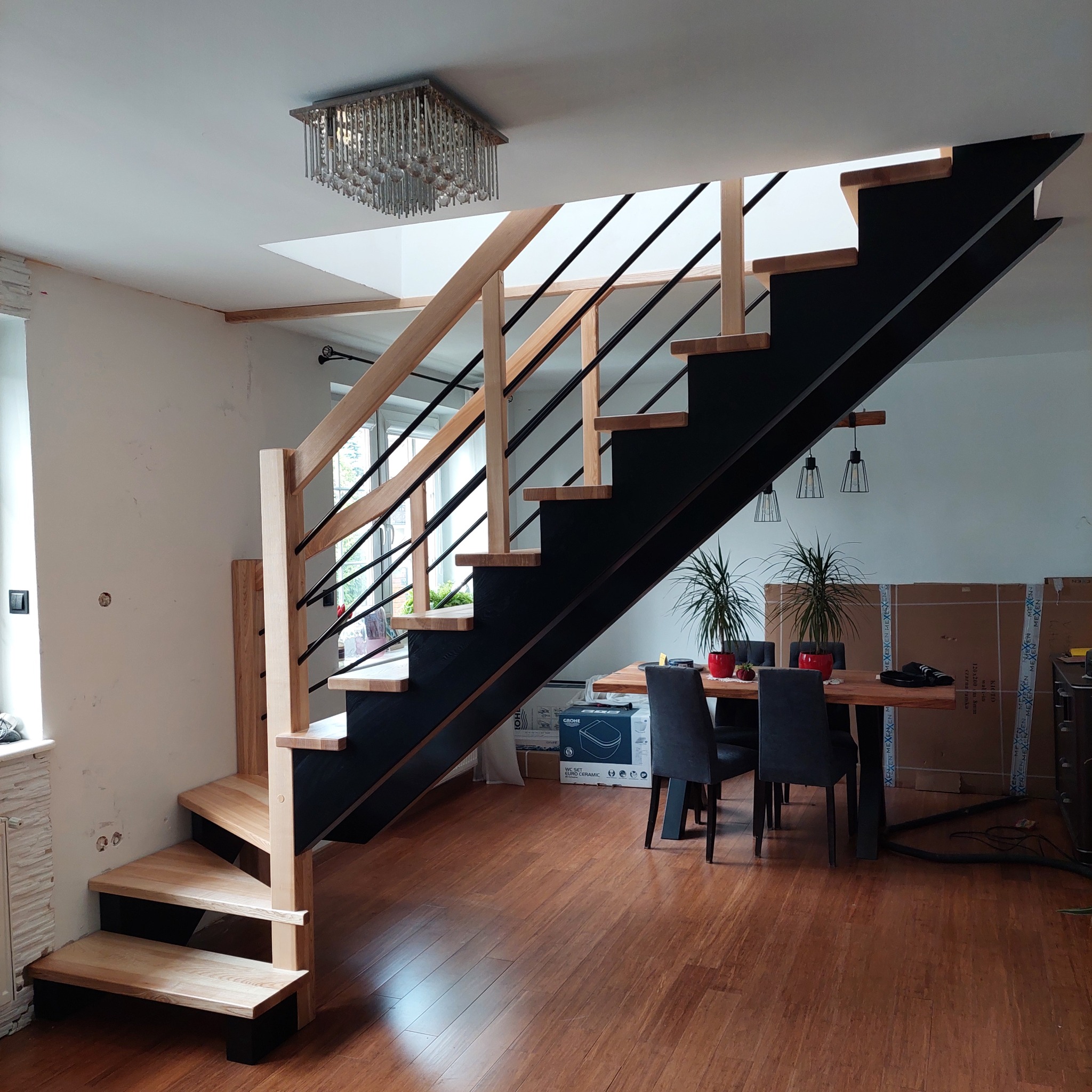 Nowoczesne schody z drewnianymi stopniami i czarną konstrukcją stalową, z balustradą z jasnego drewna i czarnymi prętami. W tle stół jadalny i kartony.