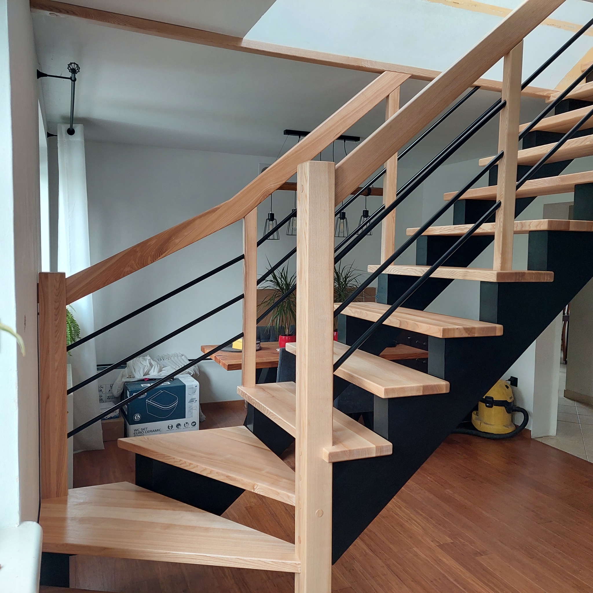 Nowoczesne schody drewniane z czarnymi elementami metalowymi i jasną balustradą, widoczne z dołu, w tle jasne wnętrze domu.