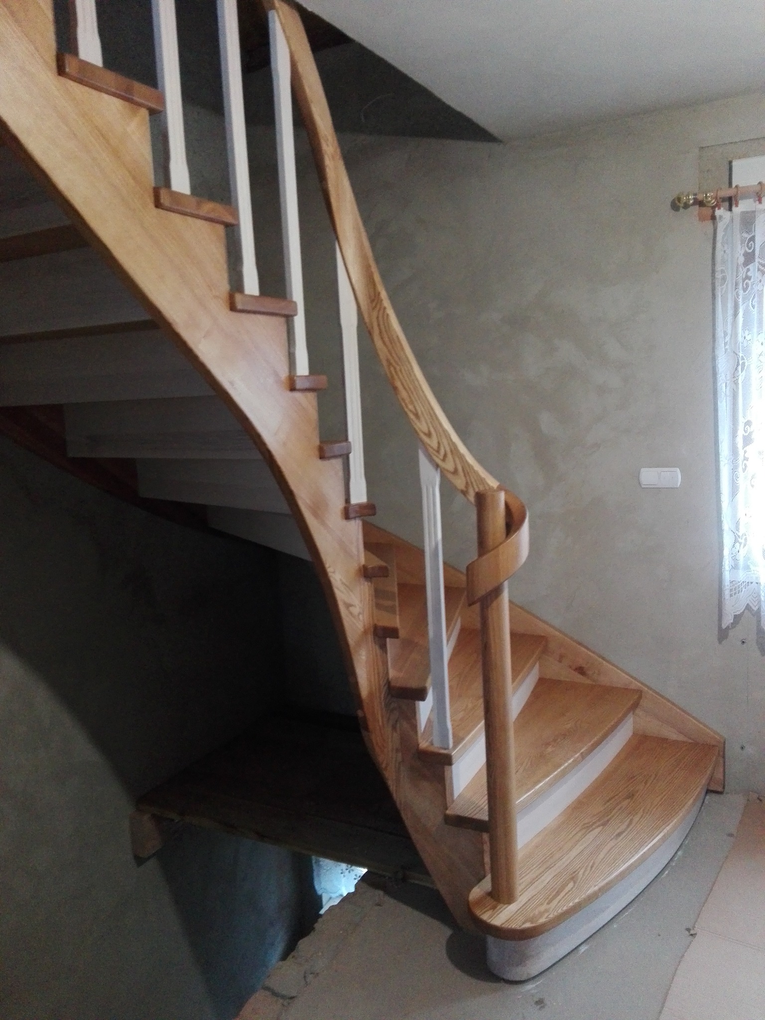 Zakręcone schody z jasnego drewna z białą balustradą, widok z dołu, naturalne oświetlenie, w tle ściana z oknem.