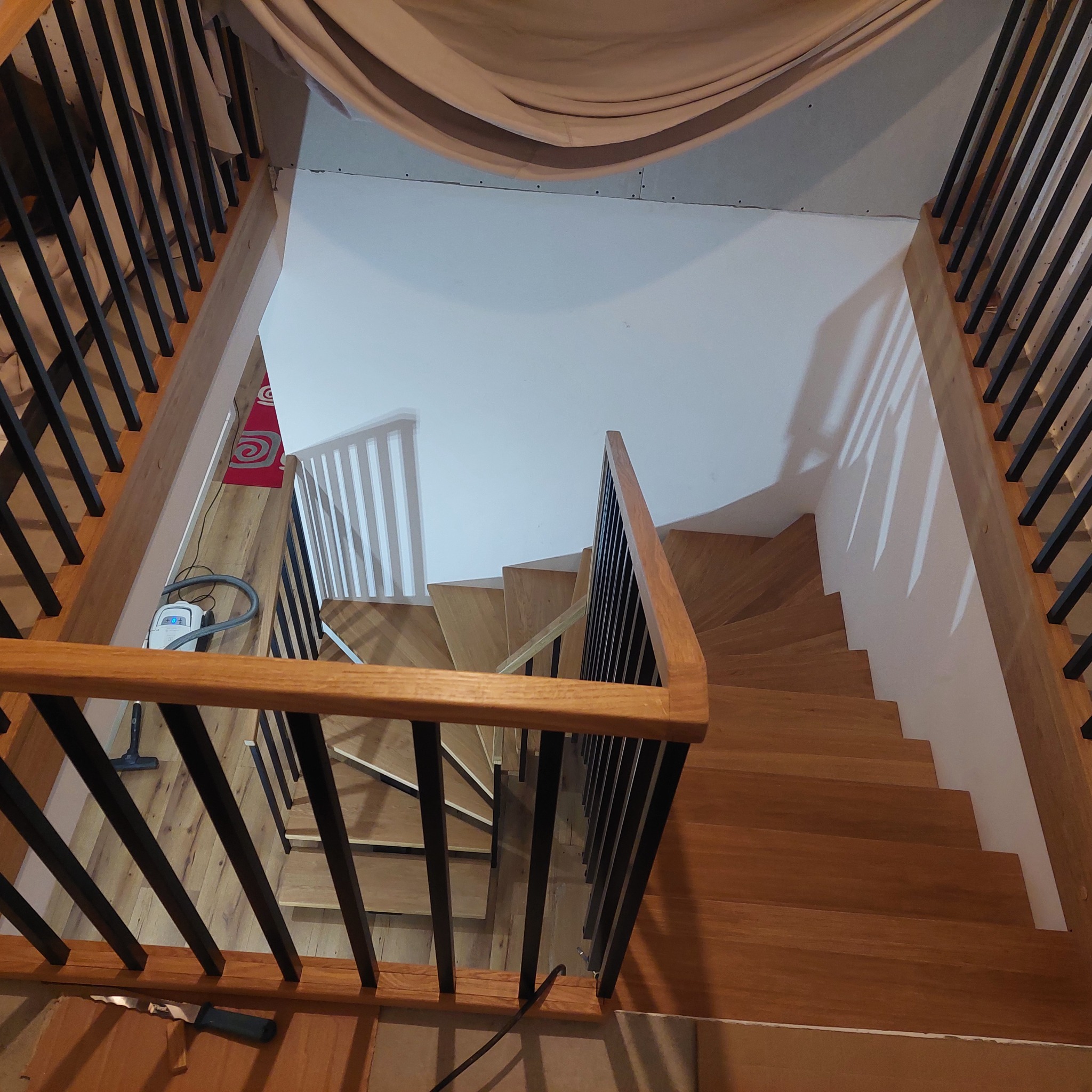 Widok z góry na drewniane schody z czarną balustradą w domu w Trzemesznie. Na podeście widoczny odkurzacz. Ujęcie z góry podkreśla spiralny kształt schodów.