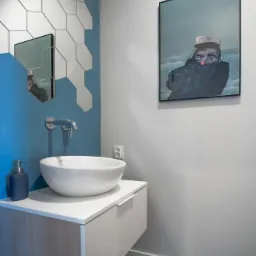 Nowoczesna łazienka z umywalką nablatową, lustrem z heksagonalnych elementów, błękitną ścianą i obrazem marynarza w tle.