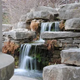 Kaskada wodna z kamieni w ogrodzie, z elementami roślinności i fragmentem kolumny po lewej stronie. Woda spływa po ułożonych blokach skalnych tworząc mały wodospad.