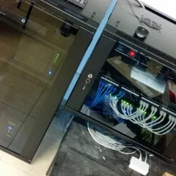 Dwie szafy serwerowe z widocznymi kablami i urządzeniami sieciowymi. Jedna szafa z kluczem w zamku. Widoczne diody LED i wentylatory chłodzące.