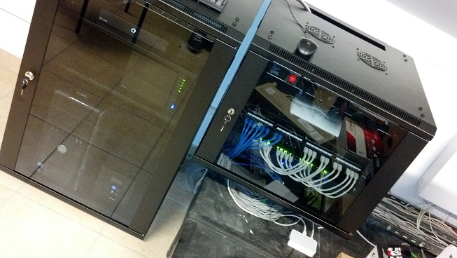 Dwie szafy serwerowe z widocznymi kablami i urządzeniami sieciowymi. Jedna szafa z kluczem w zamku. Widoczne diody LED i wentylatory chłodzące.