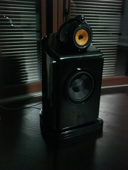 Czarny głośnik podłogowy z żółtym głośnikiem wysokotonowym na tle okna z żaluzjami. Widoczny kabel zasilający. Gładka, lakierowana obudowa.