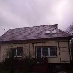 Dom jednorodzinny z nowym, ciemnoczerwonym dachem z dachówką metalową, widoczne okna dachowe i rusztowanie wokół elewacji, drabina oparta o ścianę, pochmurne niebo.