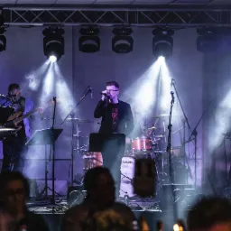 Zespół muzyczny na scenie podczas występu, wokalista z mikrofonem w centrum, perkusja Yamaha w tle, oświetlenie sceniczne i lekki dym, widoczni goście w pierwszym planie.