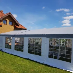 Biały namiot imprezowy z szarym dachem, ustawiony na trawniku przed domem z czerwoną dachówką, widok z zewnątrz przez okna na ustawione stoły i krzesła.