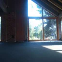 Wnętrze w stanie surowym z wylaną, ciemną posadzką, duże okna z widokiem na zieleń, ściany z czerwonej cegły dziurawki, drewniany strop.