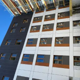 Nowoczesna fasada budynku z panelami w kolorze grafitowym, białym i szarym, z drewnopodobnymi elementami pod oknami, widoczna z dołu pod kątem, z fragmentem rusztowania na górze.