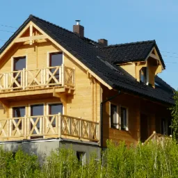 Dwukondygnacyjny dom z bali z dwoma balkonami z drewnianą balustradą w kształcie krzyży, czarną dachówką i kominem, otoczony zielenią w słoneczny dzień.