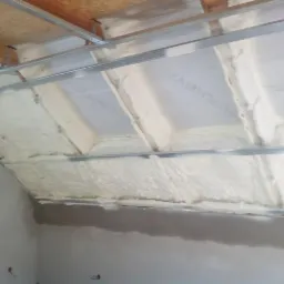 Poddasze z widoczną konstrukcją metalową i białą pianą izolacyjną, aplikowaną między krokwiami, z fragmentem instalacji elektrycznej zwisającej z sufitu.